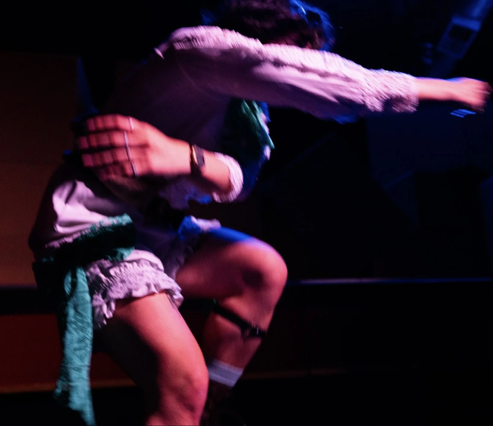 A performer in a white ruffled costume with green accents lunges forward under pink and blue stage lighting. Their body is captured in motion with one arm outstretched and the other wrapped across their torso, creating a dynamic, blurred silhouette against a dark theatrical background.