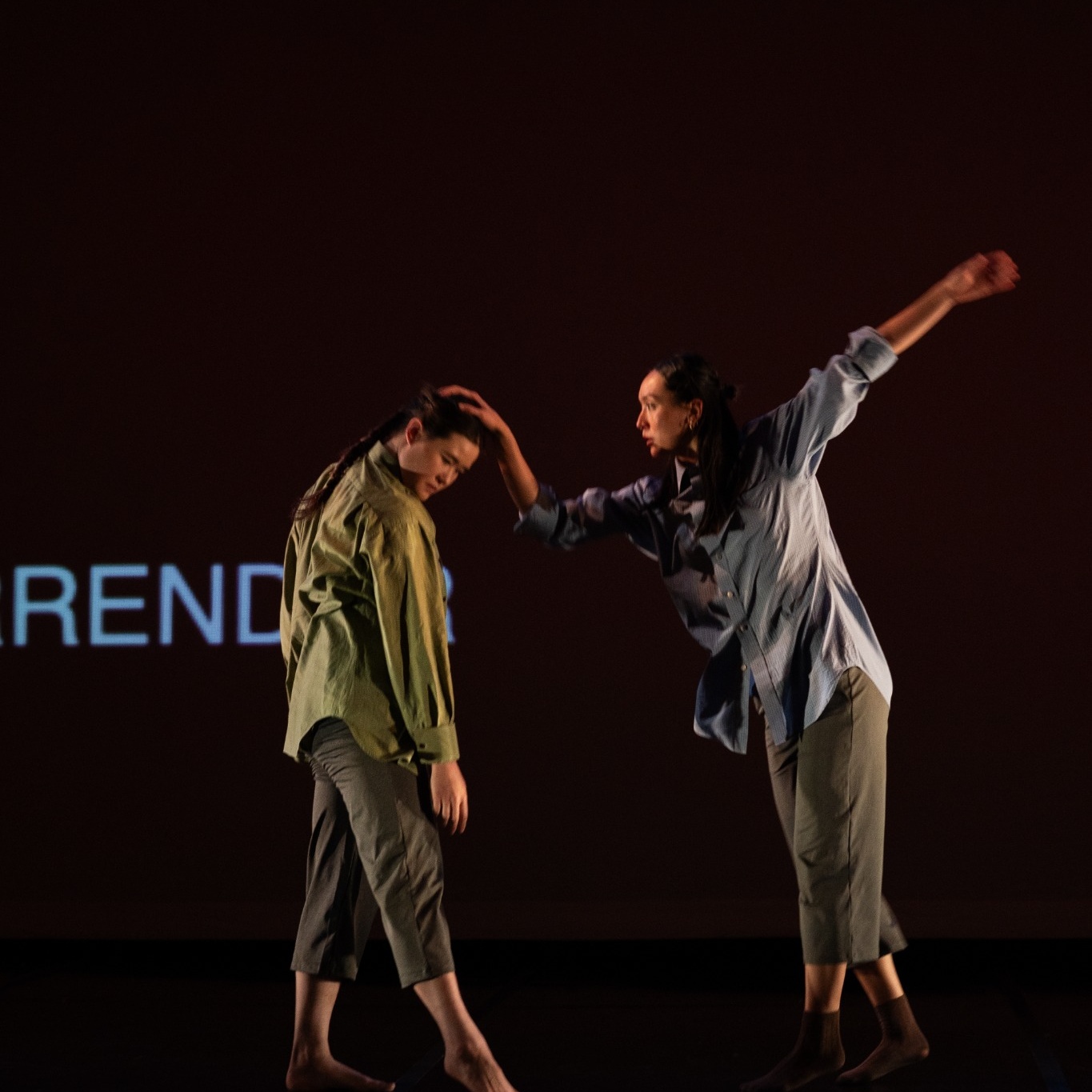 A contemporary dance performance on a dark stage. Two barefoot dancers face one another. The dancer on the left stands with their head bowed and shoulders slumped, while the dancer on the right reaches out to gently cradle the other's head with one hand. Their other arm extends outward, creating a sense of balance and openness. Both wear loose, muted button-down shirts and cropped gray pants. Warm stage lighting highlights the intimate interaction against a mostly black backdrop, emphasizing a quiet moment of comfort, care, and human connection.