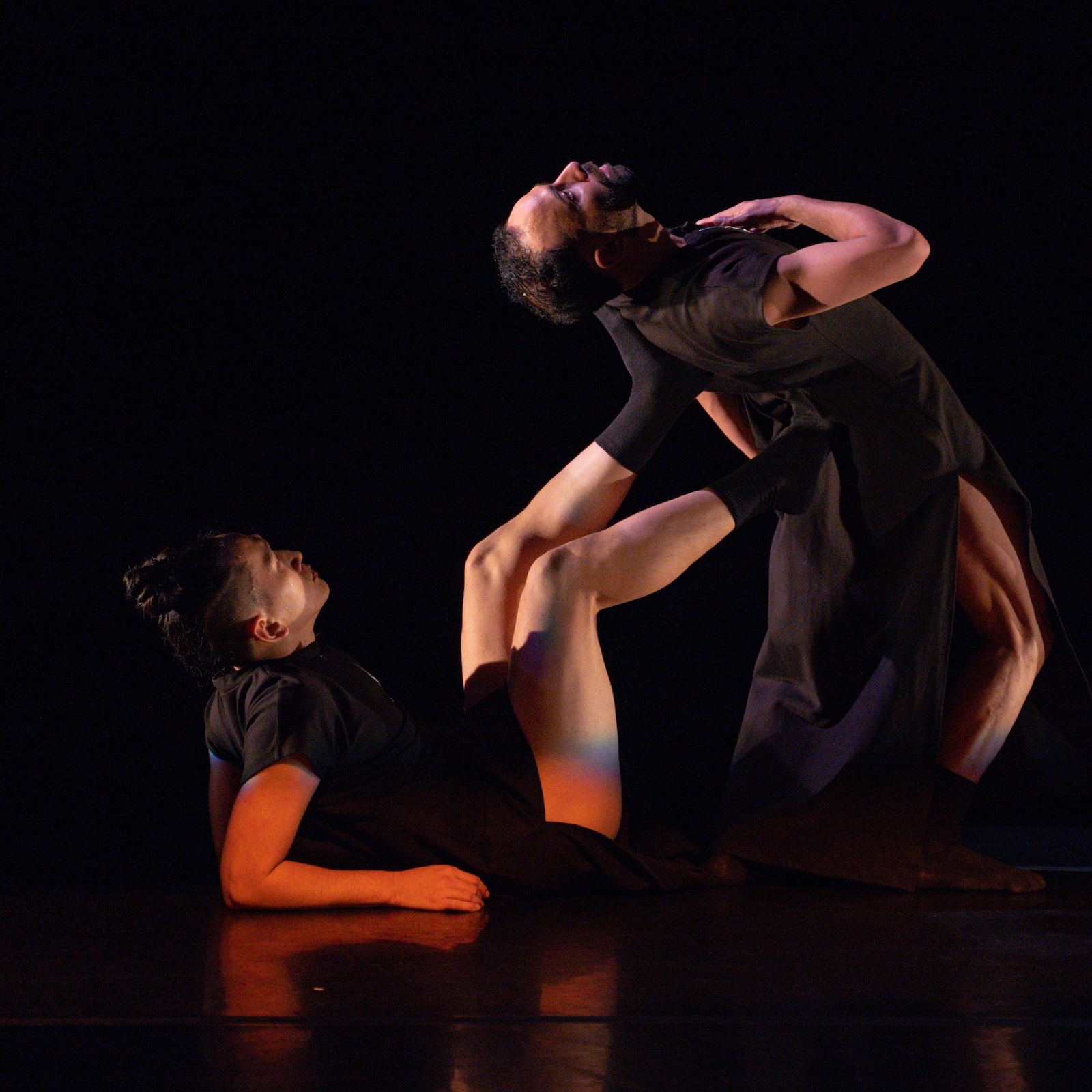 Two dancers in dark costumes against a black background. The dancer on the right is leaning backward. The dancer on the left is lying on his back, resting on his elbows, with his feet extended to support the other dancer's back.