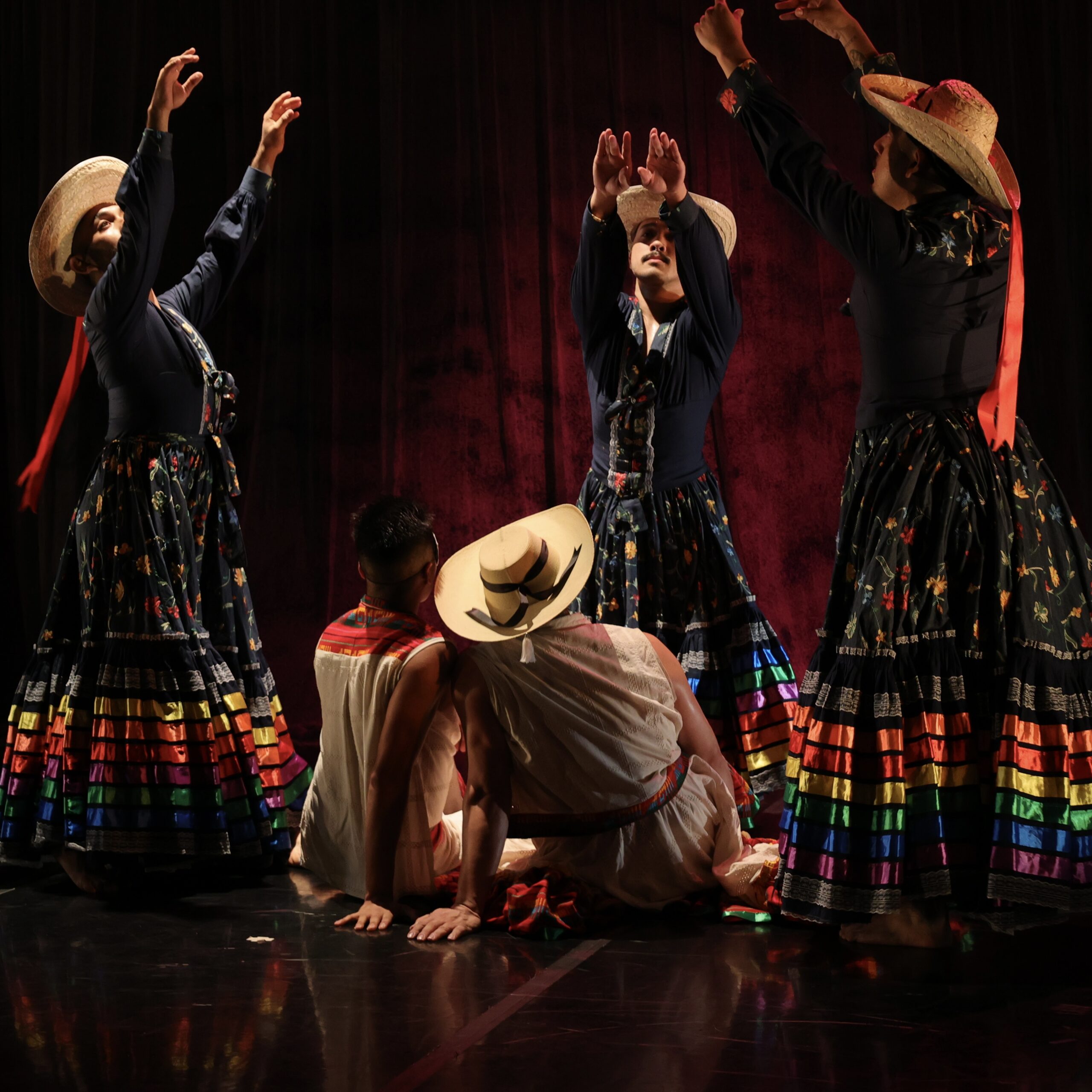 On a darkly lit stage, there are 5 dancers. Two are seated on the ground, facing back. Three surround them, standing. 