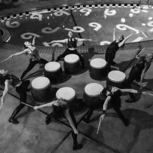 Group of 7 drummers stand in a circle looking up with arms outstretched 