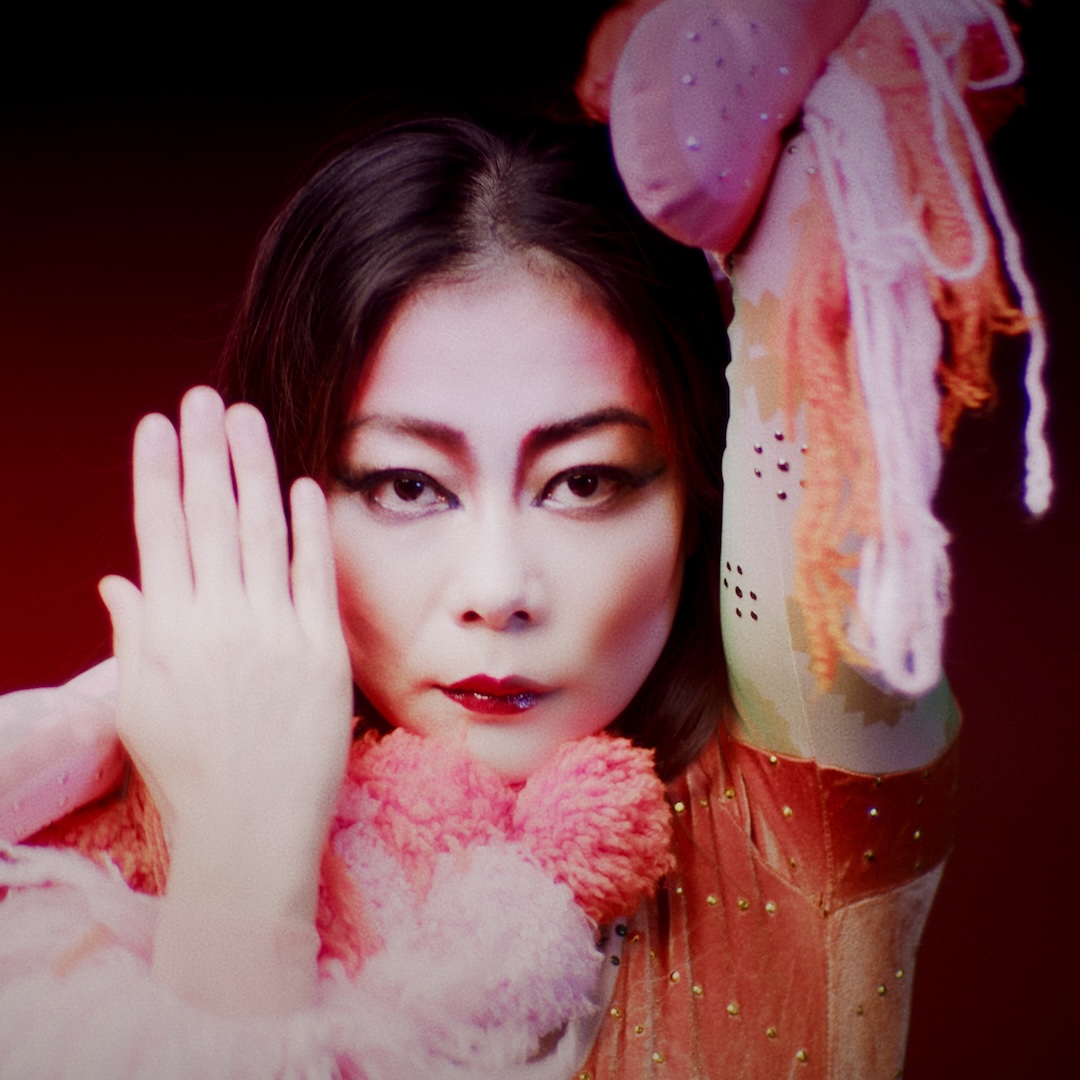 A close-up portrait of a performer gazing directly at the camera, wih dramatic mapeup, deep reed lipstick, and deep cheebones emphasized by contouring. One hand is raised to the side of their face, while the other reaches out overhead, displaying their shimmering outfit adorned with soft pinks and oranges, fluffy textures and fabrics, against a deep red backdrop. 