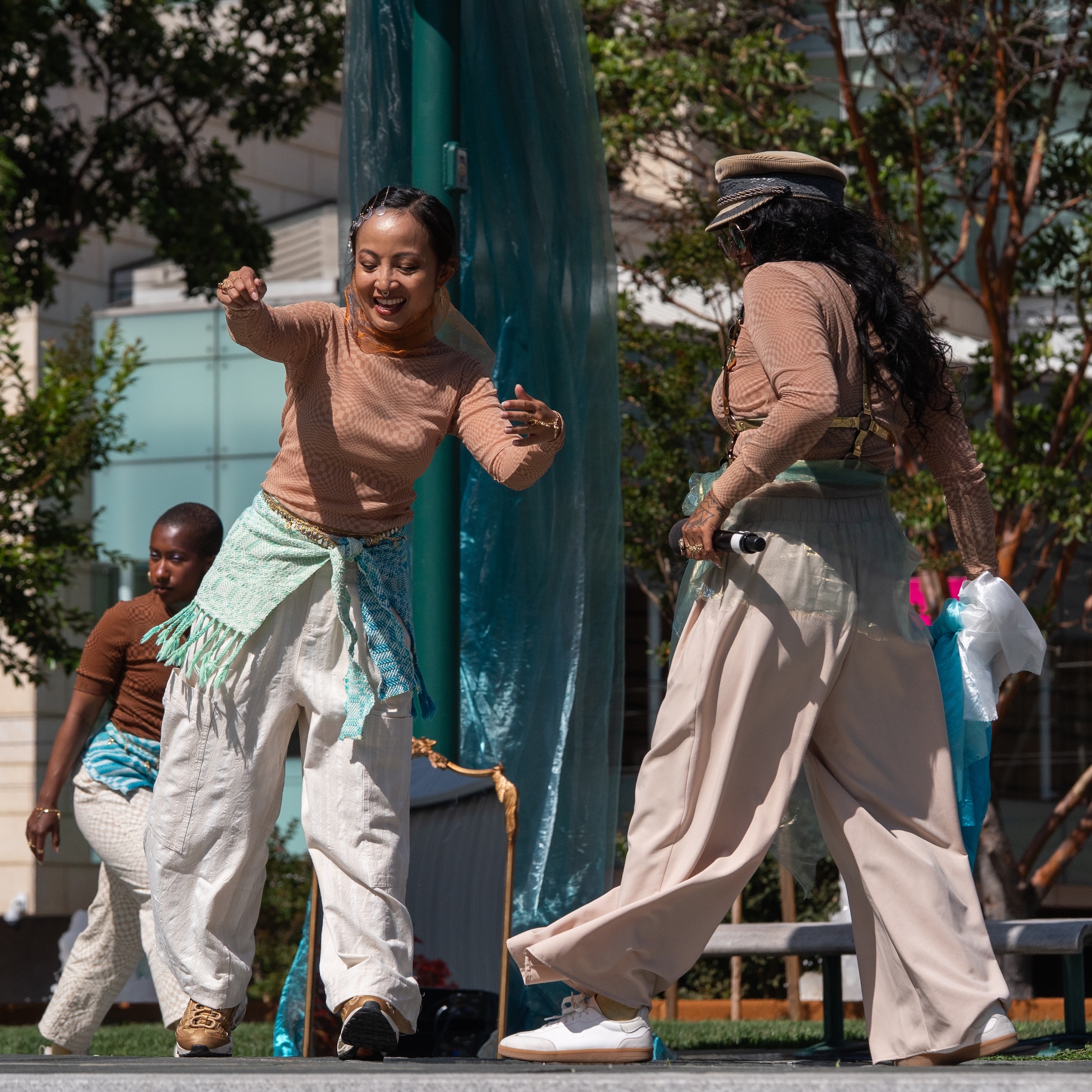 Queer, femme, and gender diverse dancers of Filipino and Black lineage wearing variations of beige, cream, and blue hues with kusikus textiles do the Filipino Cha Cha at Yerba Buena Gardens.