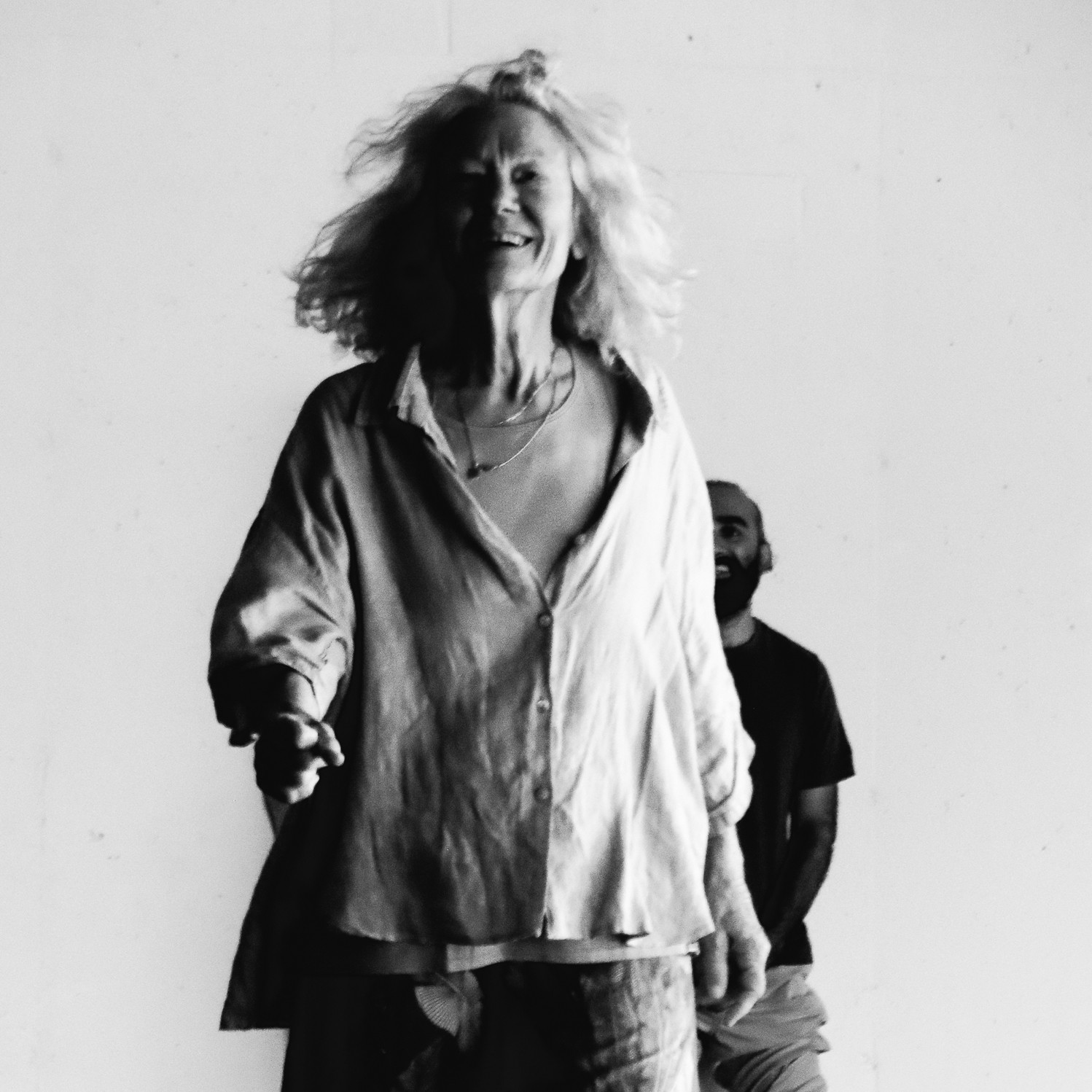 Black and white image of older woman with white hair walking towards the camera smiling with wind blowing through her hair and a loose button down shirt